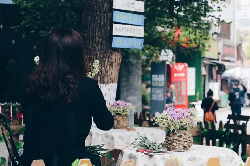 一名背对镜头的长发黑衣女子坐在街头桌前,桌上摆放着两篮淡紫色鲜花,背景是绿树和模糊的街道。