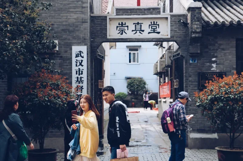 一座青砖建筑入口前,几名路人正在活动,有人在拍摄手机,有人背着背包穿行。