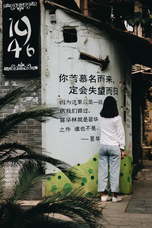 一名穿白色上衣和浅色牛仔裤的女子背对镜头,站在一面写着文字的灰墙前观看。女子身旁有绿色的植物叶片,墙上挂着复古喇叭和黑白招牌。