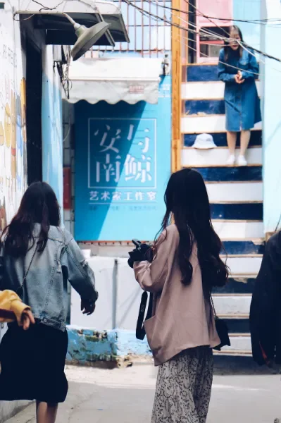 几位年轻女性背对镜头，行走在武汉的一条涂鸦巷弄中，望向远处身穿蓝色长裙登上阶梯的女子。