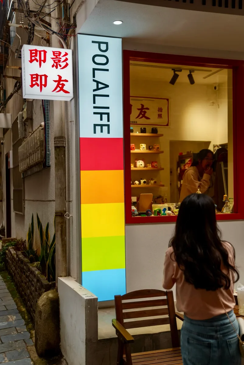 在夜晚的巷口，一位长发女子背对着镜头站在窗边。商店的招牌上写着POLALIFE和中文，橱窗内摆放着货架。透过带有红色边框的玻璃窗，可以看到店内还有另一位长发女子正在看镜子或向外观望。
