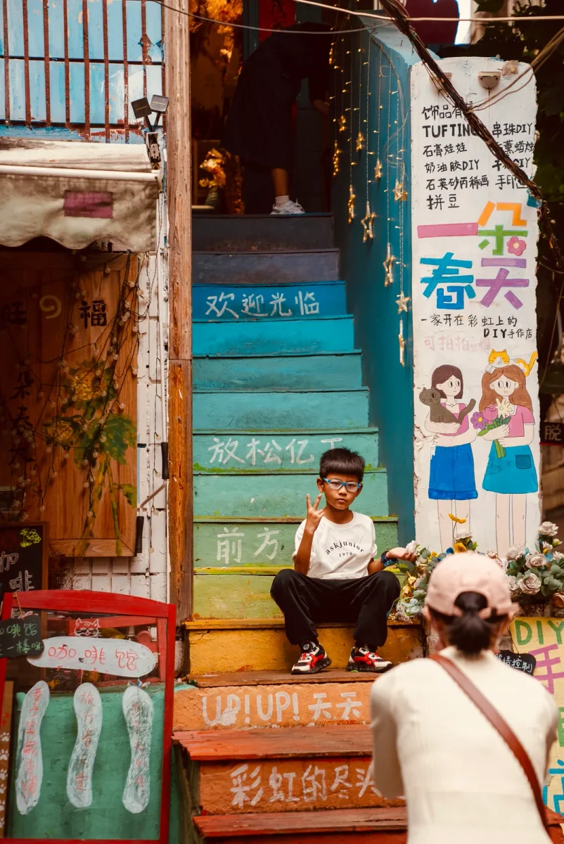 一名戴眼镜的男孩坐在彩色台阶上比出剪刀手，前方有一位戴着粉色帽子的女性正举着手机为他拍照。背景是充满涂鸦和标语的店铺入口，右侧立牌绘有卡通人物。