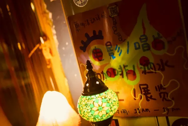 画面前景是一个发光的绿黑马赛克灯罩,背景是一张手写海报与模糊的室内角落。
