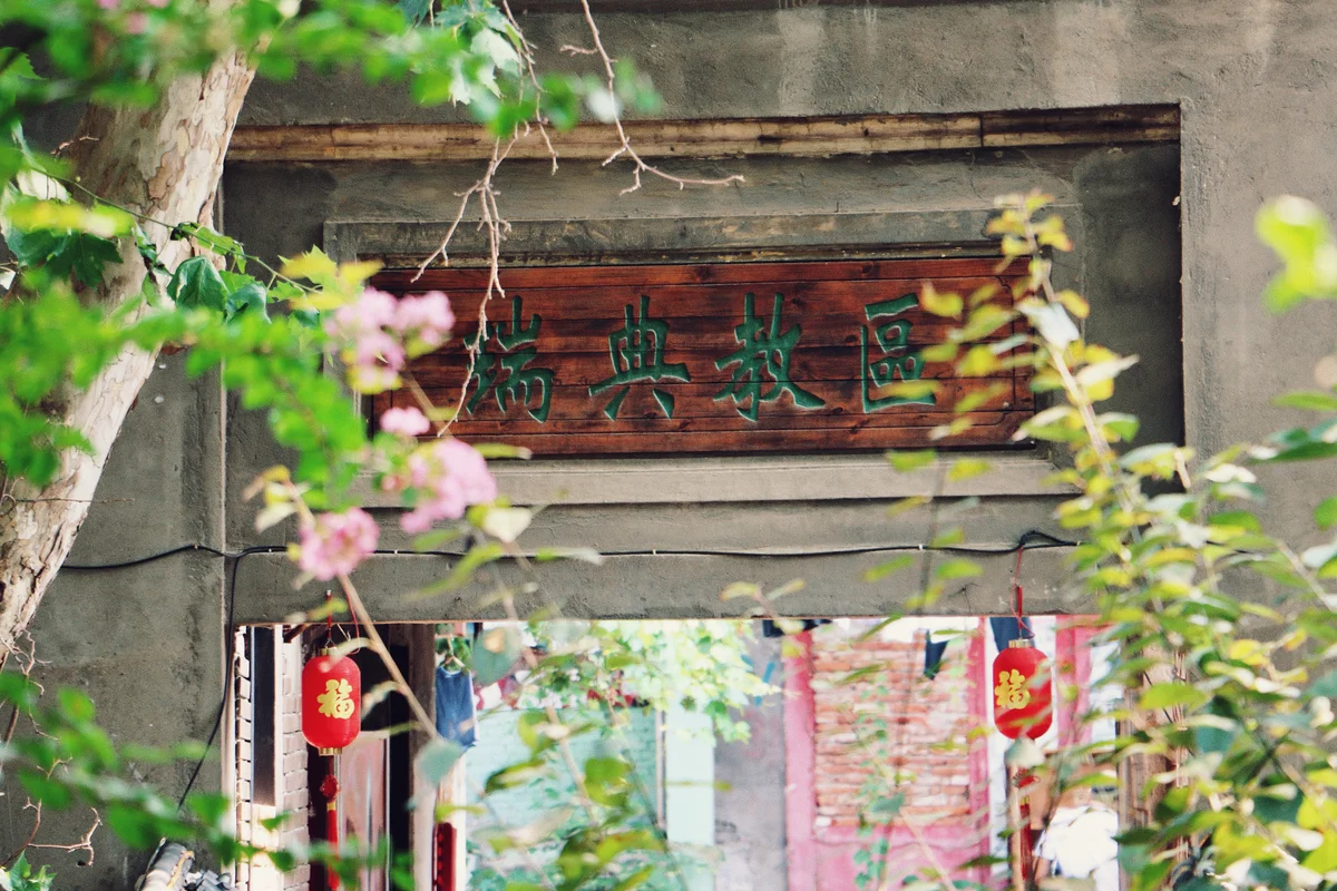 透过前景的树枝、绿叶与粉色花朵，可见一扇灰墙门洞，门楣上挂着一块写有绿色字体的深色木匾，门洞下方垂挂着两只红色灯笼，灯笼上印有金色汉字，门内可见砖墙与蓝色、白色的物件。