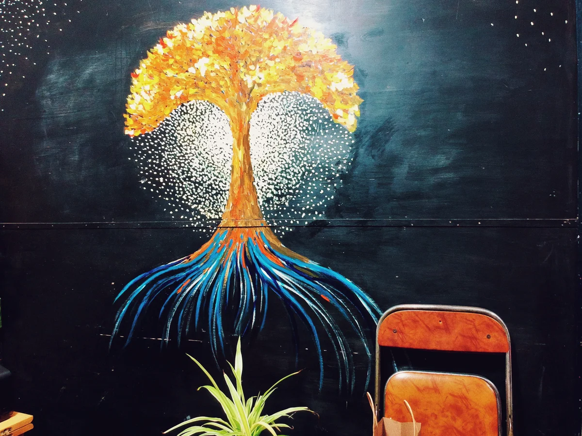 黑色墙面上一幅手绘壁画，画中一棵树的树冠为金黄，树根为蓝色，向上如扇面展开，树冠周围布满白色光点；墙前左侧有一盆绿植，右侧立着一把橙色靠背椅。