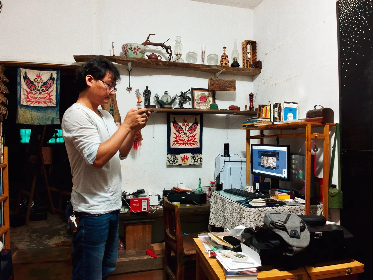 一位戴眼镜的男子站在堆满旧器物和手工艺品的房间里，双手在胸前捧着一件小物低头端详，身后木架上摆着陶罐、摆件和悬挂的织物，右侧是亮着屏幕的电脑桌。